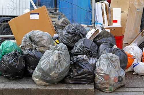 A collection of black and grey rubbish bags filled with waste material, piled on a paved pavement area outside a building or service site. The bags are made of plastic and appear to be tied at the top, with some showing slight bulges indicating contents inside. Among the bags, there are also some cardboard boxes, one large open box leaning against the pile, and a few smaller items such as plastic bottles and paper, suggestive of household or general waste. The background includes a wooden pallet, a red container, and various stored items indicating a refuse collection or clearance scene typical of rubbish removal services in areas like Beaconsfield. The scene is outdoors, with natural daylight highlighting the textures of the plastic bags and the surrounding environment, consistent with an urban or residential setting. The image reflects a typical rubbish clearance situation managed by companies like House Clearance Beaconsfield in the local postcode area.