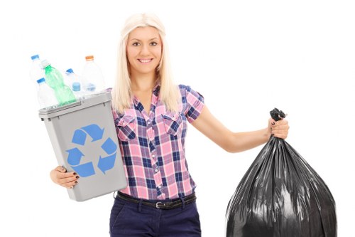 A young woman with long blonde hair, smiling and wearing a short-sleeved pink and blue checkered shirt, is standing against a plain white background. She holds a light grey recycling bin in her left hand, which is filled with various plastic bottles and containers of different colors, including clear, green, and blue. In her right hand, she grasps a tightly tied black rubbish bag. The image appears to be a promotional or illustrative photograph related to waste collection or rubbish removal services. The plain background and bright lighting highlight the objects she is holding, emphasizing the concepts of recycling and waste disposal relevant to services such as those offered by House Clearance Beaconsfield, serving the Beaconsfield area. The visual presentation is clean, neutral, and professional.