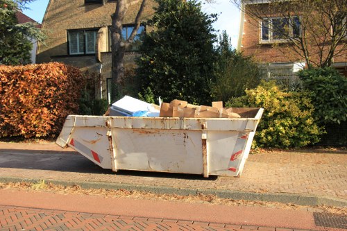 A large, rectangular metal skip with a slightly rusted and weathered white finish is positioned on a paved driveway beside a residential street in a suburban area. The skip is filled with various discarded materials, including several cardboard boxes and other unspecified waste. Surrounding the skip are some green bushes, a neatly maintained hedge with reddish-brown autumn foliage on the left, and a mix of deciduous and evergreen trees providing partial shade. In the background, there are two multi-storey houses with brick and stone facades, one with a gabled roof and the other with flat sections, typical of suburban housing. The scene is illuminated by natural daylight, with subtle shadows cast by the foliage and buildings. This image aligns with the rubbish removal services offered by House Clearance Beaconsfield, showcasing typical waste disposal in a residential setting near Beaconsfield, Bucks, highlighting the process of clearing household waste from properties.