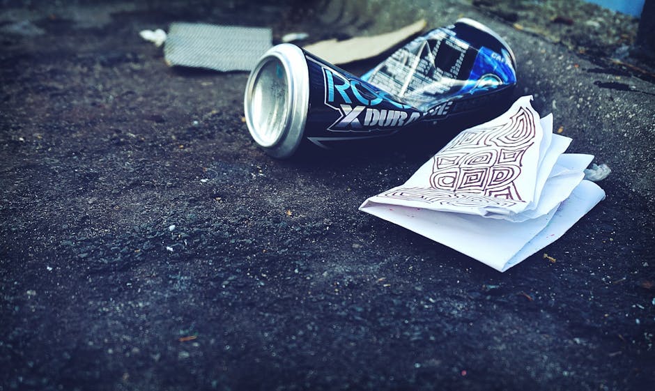 A close-up view of a discarded crushed aluminum can with a dark blue and silver design, lying on a rough asphalt surface. Next to the can, there is a small stack of white paper or napkins, partially c