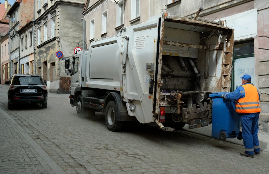 A worker standing on a paved roadside next to a large red rubbish collection truck, which is part of a waste management service often used for private waste collection or on-site clearance. The worker