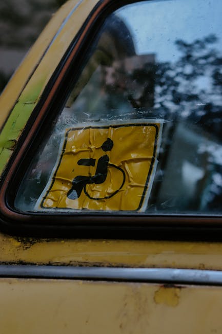 Close-up of the rear side window of a yellow vehicle, featuring a yellow disability access sticker with a black icon of a person in a wheelchair. The sticker appears wrinkled and slightly creased, wit