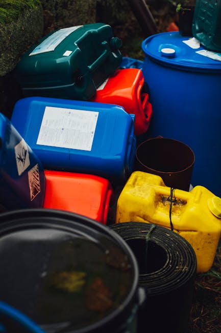 Five black wheeled rubbish bins with yellow lids lined up along a grey concrete ground in front of a plain white wall. Each bin has a white oval label with identification numbers, and they are positio